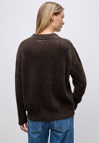 Maglione marrone e morbido con un ampio colletto e una vestibilità rilassata, abbinato a jeans di denim blu. La texture sembra soffice e calda.