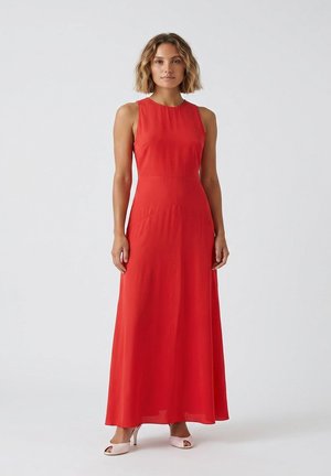 Donna in piedi che indossa un abito rosso senza maniche lungo fino a terra e décolleté rosa chiaro con punta aperta su uno sfondo chiaro e uniforme.