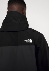 Personne vue de dos portant une veste The North Face noire et gris foncé avec une capuche, logo visible sur le haut du dos.