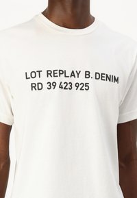 Balta kokvilnas T-kreklā ar uzrakstu treknām melnām burtiem: "LOT REPLAY B. DENIM RD 39 423 925." Arapa, īsās piedurknes, standarta piegriezums.