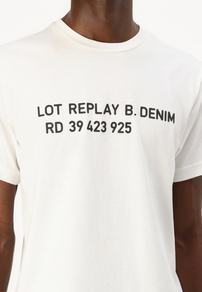 T-shirt en coton blanc avec un texte noir audacieux : "LOT REPLAY B. DENIM RD 39 423 925." Col rond, manches courtes, coupe standard.