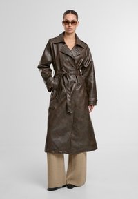 Brązowy płaszcz trench z sztucznej skóry, długi, z paskiem w talii, z podwójnymi guzikami i szerokim kołnierzem. Modelka nosi szerokie beżowe dzianinowe spodnie.