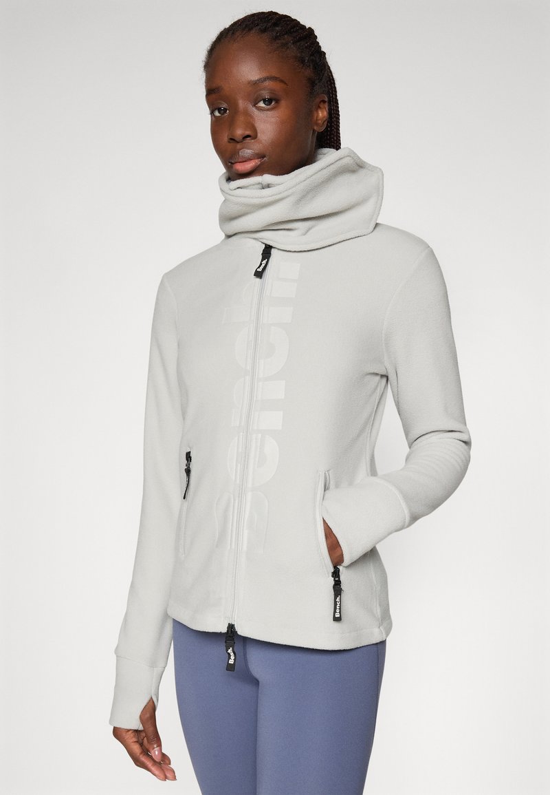 Vrouw in een lichtgrijze fleece jack met ritssluiting en hoge kraag, met duimgaten, gecombineerd met blauwe leggings, staand tegen een effen achtergrond.