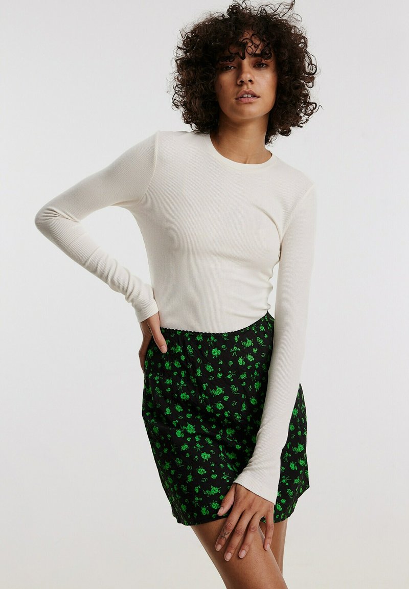 Crème ribbeltrui met lange mouwen, gecombineerd met een zwarte rok met een groen bloemenpatroon. De top heeft aansluitende mouwen en een ronde halslijn.