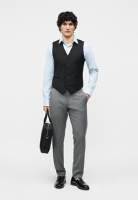 Νέος άνδρας που φοράει μαύρο γιλέκο, ανοιχτό μπλε πουκάμισο, γκρι παντελόνι, μαύρα παπούτσια, κρατώντας μια μαύρη τσάντα, στέκεται μπροστά σε απλό φόντο.