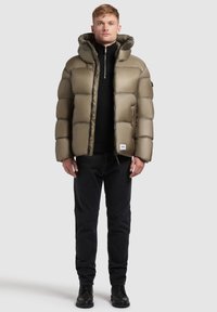 Padded olive green jacket with a hood, zip closure, and side pockets. Worn over a black sweater and black pants, paired with black boots.