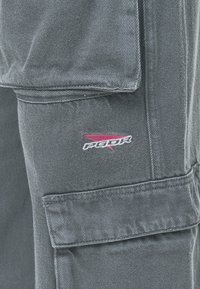 Calças cargo cinzas com um tecido texturizado, costuras visíveis e um pequeno logótipo gráfico em vermelho e preto no bolso.
