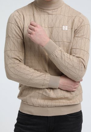Pull beige à col roulé avec motif en losange porté par une personne les bras croisés, accompagné d'un pantalon noir.