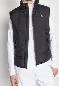 Schwarze ärmellose Steppweste mit hohem Kragen, Frontreißverschluss, zwei Seitentaschen und dezent strukturiertem Stoff; Logo auf der oberen linken Brust.