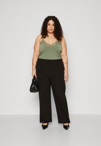 Grünes Spaghetti-Trägershirt mit Spitze, kombiniert mit schwarzen weit geschnittenen Hosen. Model hält eine kleine schwarze Tasche und trägt schwarze High Heels.