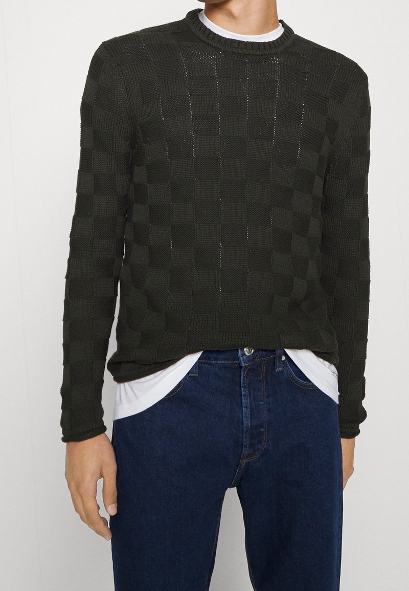 Donkergroene gebreide trui met een geometrisch patroon, ronde halslijn en lange mouwen, gedragen over een wit hemd en gecombineerd met blauwe jeans.