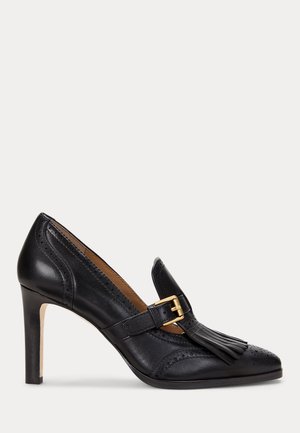 Chaussure habillée à talon haut en cuir noir avec perforations décoratives, frange avant et bride boucle dorée sur fond blanc.