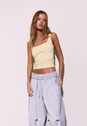 Giovane donna con capelli lunghi che indossa un top asimmetrico color giallo pallido e pantaloni della tuta larghi grigio chiaro, davanti a una parete chiara e semplice.