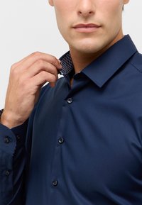 Navyblauw overhemd met een subtiele glans, voorzien van een patroon in de binnenkraag en manchetten, en plastic knopen aan de voorkant.