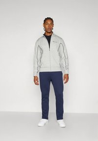 Pelēka džemperis ar rāvējslēdzēju un melnu apmali, kombinēts ar tumši ziliem sporta biksēm. Modelis valkā baltas sporta kurpes un zem džempera melnu T-kreklu.