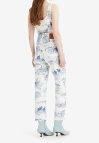 Overoles de mezclilla azul y blanco con un cuerpo ajustado y piernas anchas, que presentan un parche de cuero marrón y botas de ante de color azul claro hasta el tobillo.