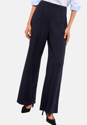 Femme portant un pantalon noir taille haute à jambes larges, associé à des talons noirs à bouts pointus et une chemise bleu clair aux manches retroussées.