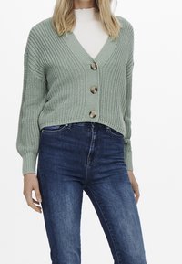 Groene gebreide cardigan met V-hals, knopen aan de voorkant en geribbelde manchetten, gecombineerd met donkerblauwe jeans. Gewatteerde stof en vier knopen.