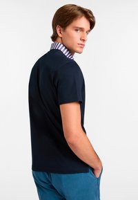 Granatowa koszulka polo z krótkim rękawem i paskowanym kołnierzykiem. Materiał wydaje się miękki, a krój jest swobodny. Noszona z jasnoniebieskimi spodniami.