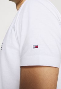 Camiseta de algodón blanca con mangas cortas; presenta un pequeño logo bordado en la manga en rojo, blanco y azul. Textura suave.