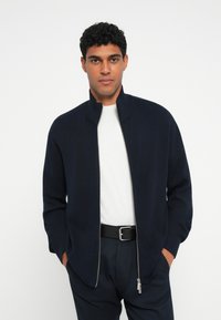 Casaco azul marinho com fecho, de colarinho alto, feito de tecido suave. Usado sobre uma camisa branca, combinado com calças escuras e um cinto preto.