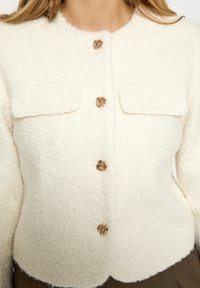 Kreminės spalvos tekstūruotas švarkas su trumpu apykakliu, dviem priekinėmis kišenėmis ir auksinės spalvos apvaliais mygtukais. Prigludęs siluetas, minkšta medžiaga su jaukia išvaizda.