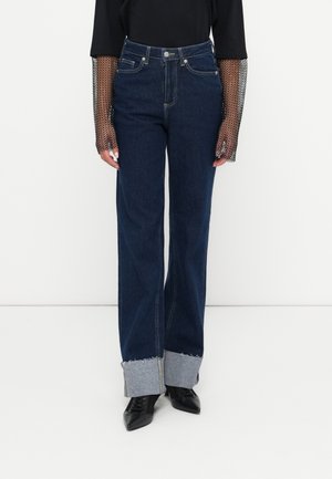Wide-leg dark blue denim jeans with a raw hem, featuring light blue cuffs and front pockets. Paired with a black top with mesh sleeves.