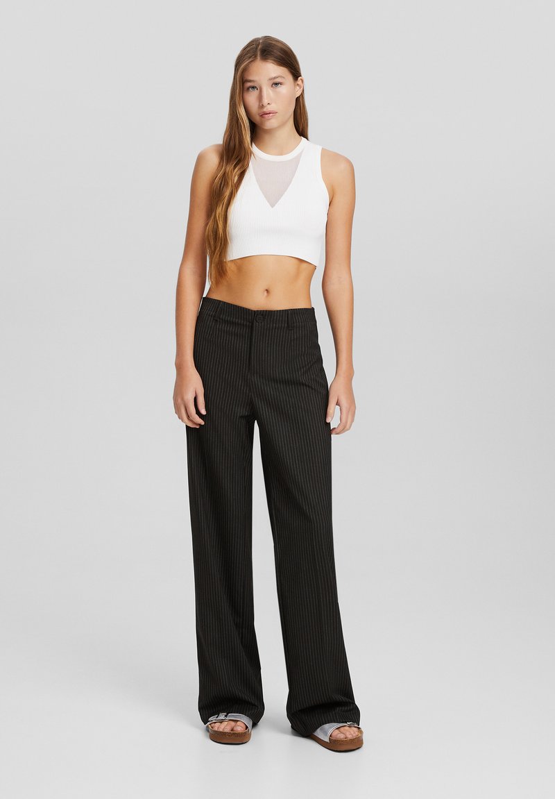 Débardeur blanc côtelé à décolleté en V associé à un pantalon large à rayures noires, comportant une fermeture à bouton sur le devant et des sandales élégantes.