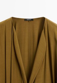 Tessuto verde oliva con un dettaglio del colletto drappeggiato, caratterizzato da una cucitura ordinata e un'etichetta con la scritta "Massimo Dutti". Morbida e liscia al tatto.