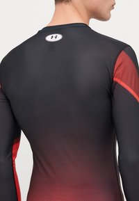 Chemise ajustée à manches longues noire avec des accents rouges, présentant un motif dégradé. Fabriquée dans un matériau lisse et extensible. Comprend un logo au dos.