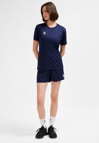 Naine seisab, käed selja taga, seljas tumesinised spordiriided, valged tavatsokid ja mustad spordijalanõud, valgel taustal.