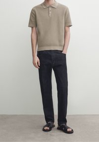 Khakinvärinen neulottu poolopaita, jossa on lyhyet hihat ja kolmen napin läpimenoaukko, yhdistetty tummansinisiin farkkuihin ja mustiin sandaleihin. Sileä tekstuuri.