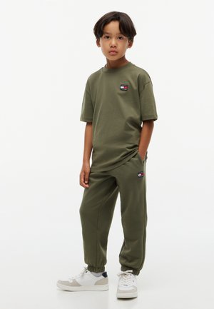Enfant debout portant un t-shirt vert olive assorti et un jogging avec des baskets blanches sur fond blanc uni.