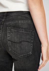 Gros plan sur une personne portant un jean en denim noir, montrant la poche arrière et une partie d'une chemise blanche ainsi qu'un bras.