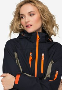 Giacca da esterno navy con zip e dettagli arancioni, dotata di cappuccio strutturato, numerose tasche con zip e tessuto texturizzato.