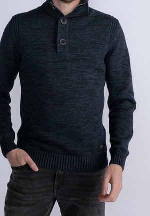 Homme portant un pull en maille bleu foncé avec poignets et col côtelés, deux boutons près du cou, associé à un jean noir.