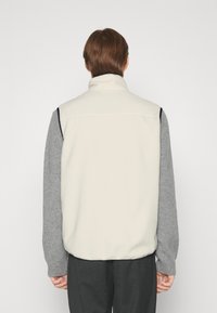 Chaleco masculino con cuello alto; frente de color crema, mangas de lana gris; textura suave; sin logotipo visible; capa de estilo con contraste tonal.