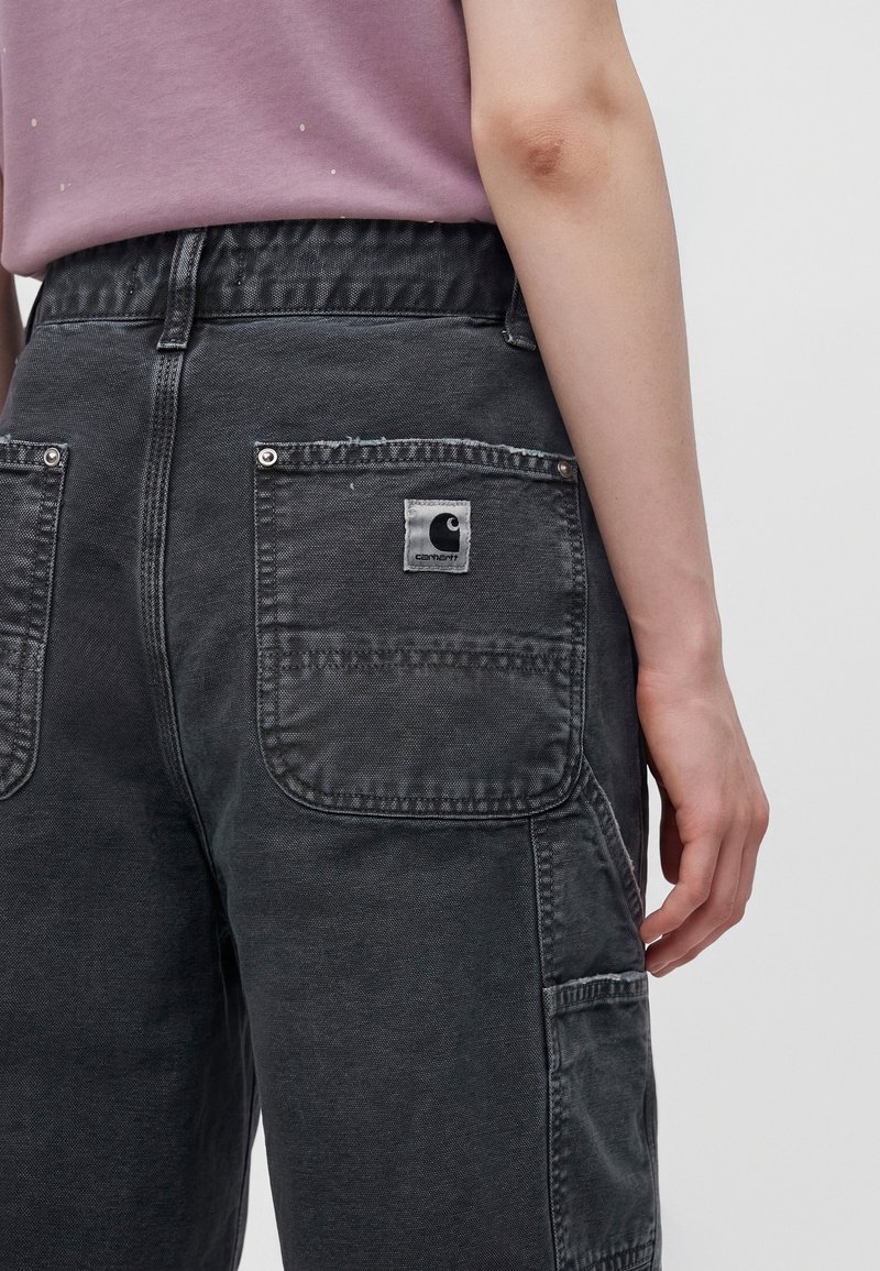 Nahaufnahme einer Person, die schwarze Carhartt-Denim-Hosen mit Gesäßtaschen und ein violettes Hemd trägt, Hand ruht seitlich.