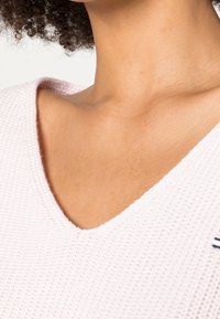 Vêtement tricoté rose clair avec un décolleté en V. Présente un tissu texturé et un petit accent marine d'un côté près de l'épaule.