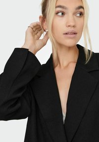 Blazer nero con una vestibilità tailored, revers affilati e maniche ampie. Il tessuto appare strutturato, indossato sopra un top chiaro, con orecchini minimalisti.