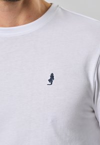 T-shirt bianco in cotone con maniche corte e scollo rotondo. Presenta una piccola figura ricamata blu marina nell'area del petto sinistro.