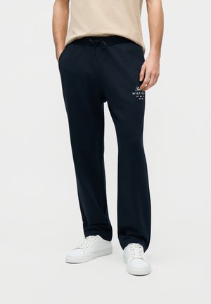 Osoba v čiernych teplákoch so šnúrkou s bielym logom Tommy Hilfiger na ľavom stehne a bielych teniskách, stojaca pred jednofarebným pozadím.