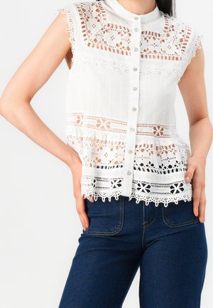 Blouse blanche sans manches avec panneaux en dentelle florale et boutonnière devant, portée avec un jean taille haute bleu foncé, mains posées sur les hanches.