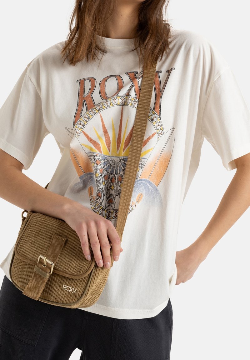 Personne portant un t-shirt blanc Roxy avec un motif soleil et planche de surf, tenant un petit sac bandoulière marron texturé sur un pantalon noir.