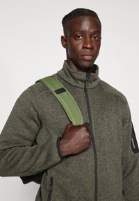 Grüne Fleecejacke mit hohem Kragen, schwarzem Reißverschluss und Reißverschlusstaschen. Hält einen grünen Riemen eines Rucksacks fest.