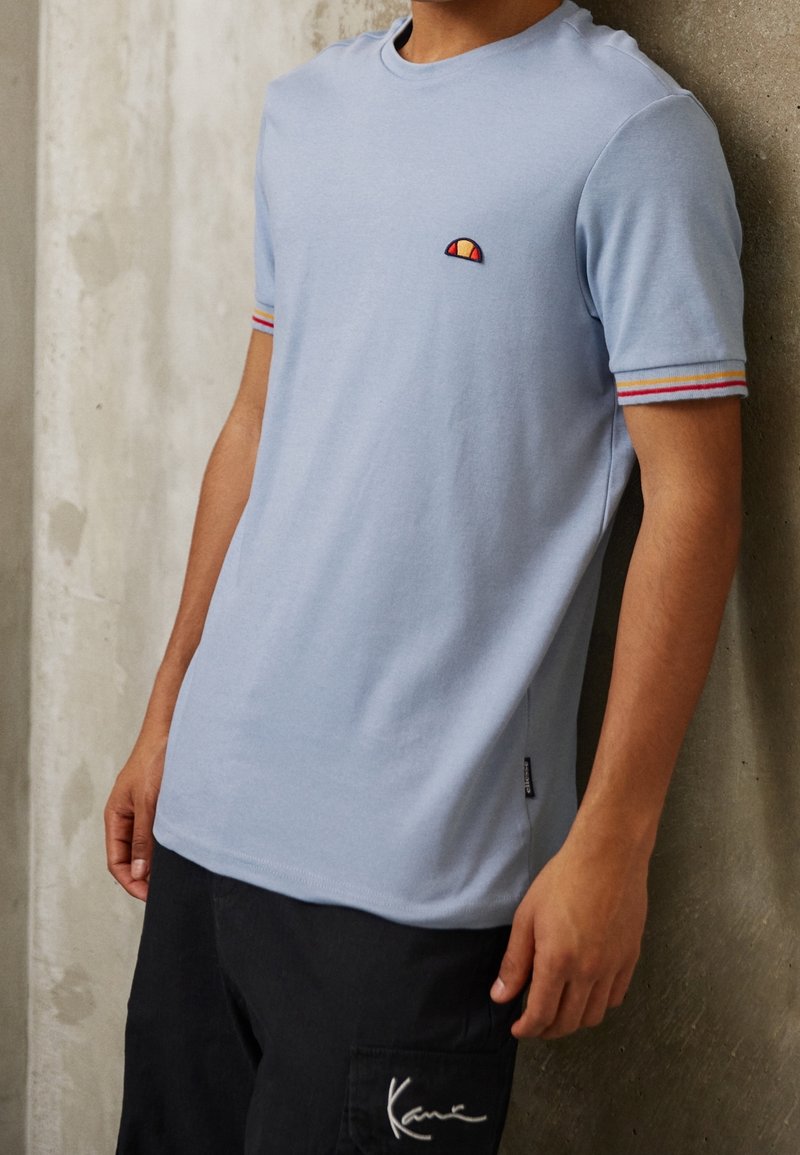 T-shirt en coton bleu clair à manches courtes, arborant un petit logo rouge et jaune ainsi que des accents rayés en rouge, jaune et bleu sur les manches.