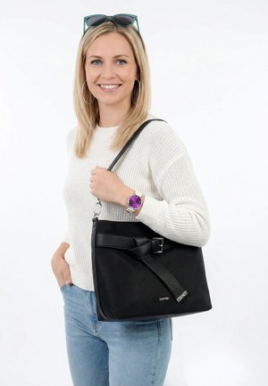 Smilende kvinde med blondt hår iført hvid sweater, blå jeans, lilla ur, solbriller på hovedet og bærer en stor sort skuldertaske med spænde.