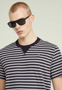 Czarne okulary przeciwsłoneczne z prostokątnymi oprawkami; model ma na sobie szary i granatowy t-shirt w paski z okrągłym dekoltem i subtelnym logo na piersi.