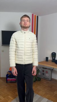 Λευκό καπιτονέ μπουφάν με ψηλό γιακά, φερμουάρ μπροστά και πλαϊνές τσέπες, σε συνδυασμό με μαύρο παντελόνι. Εσωτερικό δωμάτιο με μίνιμαλ διακόσμηση.