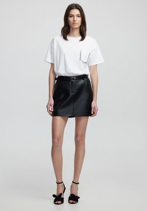 T-shirt blanc avec un détail de poche en strass, associé à une mini jupe en cuir noir et des talons noirs avec des accents de nœud.
